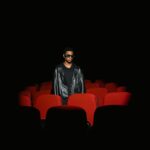 Man wearing 3D glasses and leather jacket stands alone in dark cinema.
