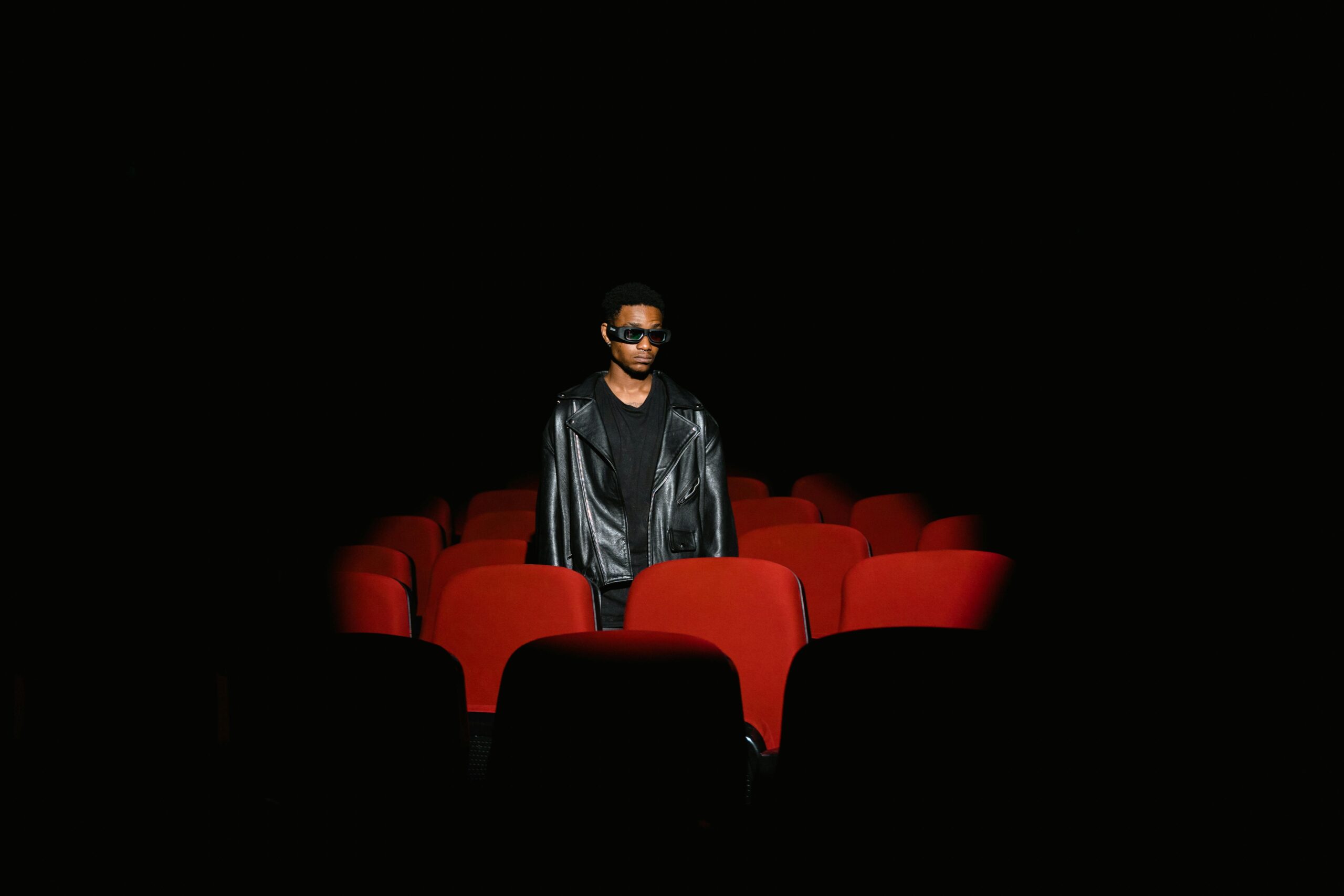 Man wearing 3D glasses and leather jacket stands alone in dark cinema.